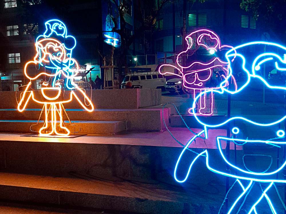 Instalação luminosa em uma praça à noite, composta por figuras de personagens coloridos feitos com luzes de neon. No primeiro plano, uma figura grande em azul e outra em rosa aparece parcialmente. Ao fundo, duas figuras completas, uma em neon amarelo e outro azul, estão posicionadas sobre degraus de concreto.