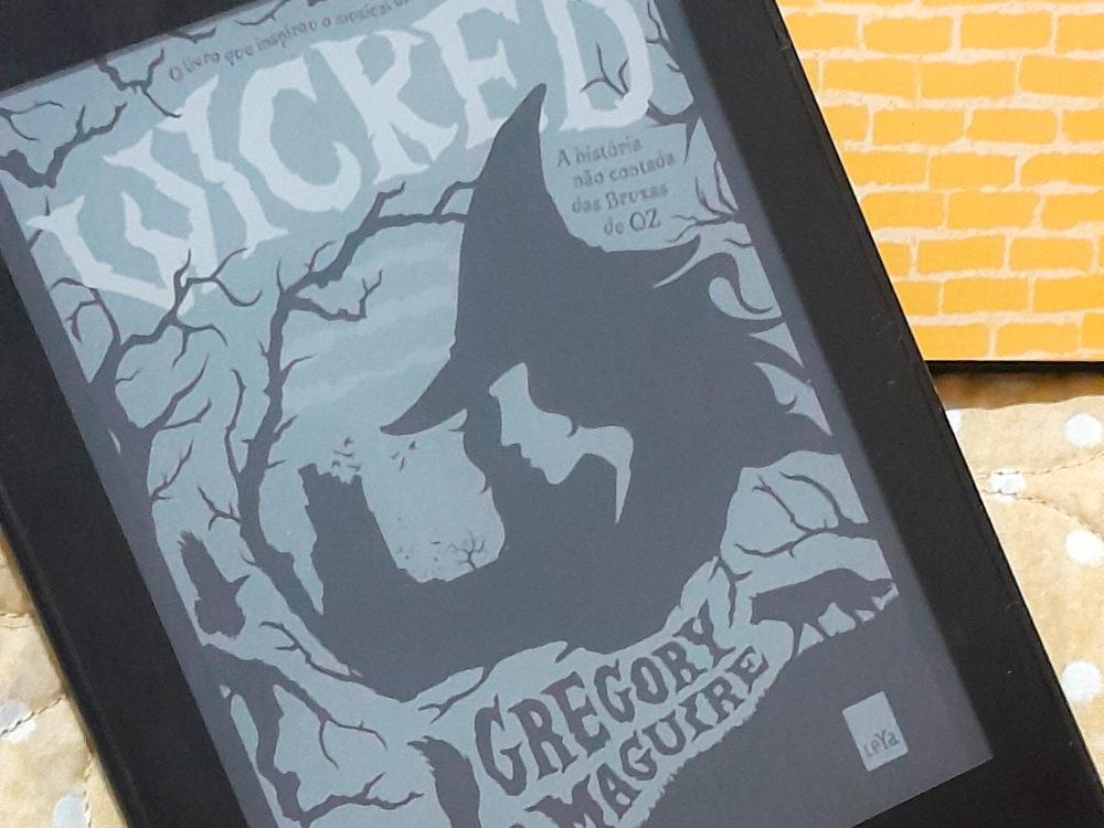 Leitor digital exibindo a capa do livro Wicked, de Gregory Maguire. A capa mostra o contorno do rosto de uma bruxa de chapéu pontudo, em meio a galhos retorcidos e formas sombrias, com o título em letras grandes na parte superior. O subtítulo diz: A história não contada das bruxas de Oz. O dispositivo está apoiado sobre um tecido claro com bolinhas e, ao fundo, há papel com estampa da estrada de tijolos amarelos.