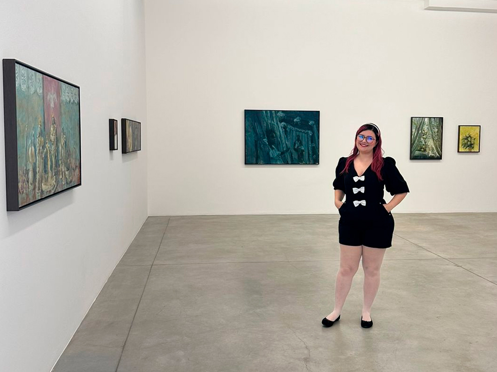 Pessoa de cabelos longos com mechas rosadas e óculos de armação marrom posando sorridente em uma galeria de arte contemporânea. Ela veste um macacão preto com três laços brancos na frente e sapatilhas pretas. Ao fundo, várias pinturas coloridas estão penduradas nas paredes brancas da exposição, com aspecto impressionista e representando imagens com flores.