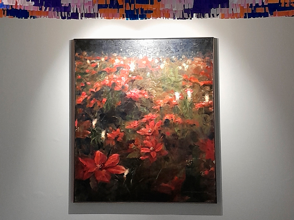 Pintura de Diego Mouro com diversas flores vermelhas vibrantes sobre um fundo escuro e esverdeado, com iluminação direcionada destacando os detalhes da obra. Na parte superior, há tiras de papel colorido em tons de roxo, laranja e branco, penduradas no teto, compondo a instalação.