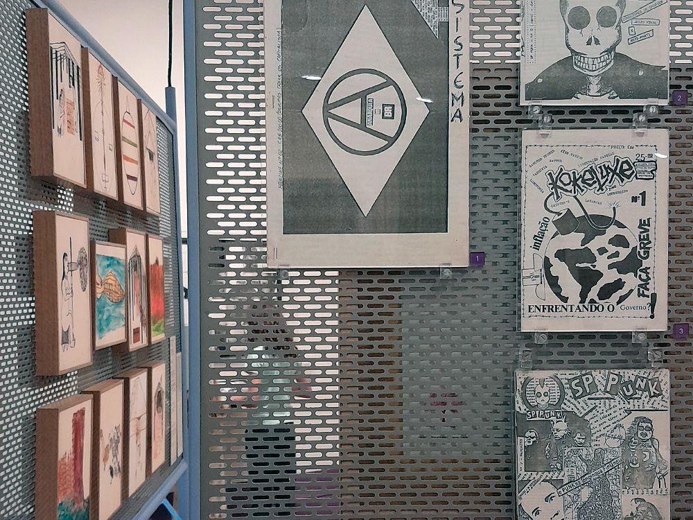 Painel de metal perfurado exibindo zines e desenhos. À esquerda, várias pequenas ilustrações coloridas emolduradas em madeira, à direita, fanzines em preto e branco com estética punk e temática política, incluindo capas com caveiras e símbolos anarquistas. Iluminação neutra e disposição organizada sugerem montagem expositiva.