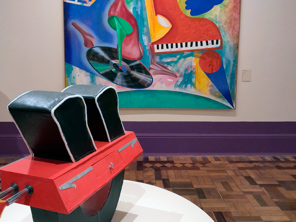 Escultura geométrica em madeira e couro preto, com base vermelha e formas que lembram um instrumento musical estilizado. Ao fundo, uma pintura colorida com piano, disco de vinil e formas abstratas em tons vibrantes de vermelho, verde e azul. O ambiente tem piso de madeira e rodapé roxo, parte da exposição Fullgás no CCBB.