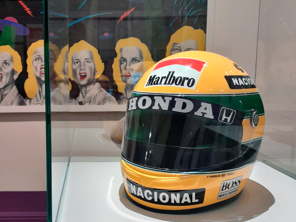 Fotografia de um capacete amarelo com faixas verdes e azuis, exibido em vitrine de vidro. O capacete tem os logotipos Marlboro, Honda, Nacional e Boss. Ao fundo, há uma obra pop art com rostos femininos em tons de cinza, cabelos amarelos e expressões de espanto.