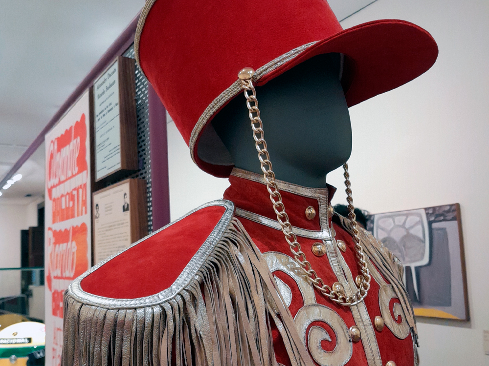 Close de um manequim vestindo um figurino vermelho de veludo com detalhes dourados, franjas nos ombros e botões dourados. O chapéu alto, também vermelho, tem aba larga e corrente dourada pendendo de um lado. O traje está exposto em ambiente museológico com painéis e obras ao fundo.