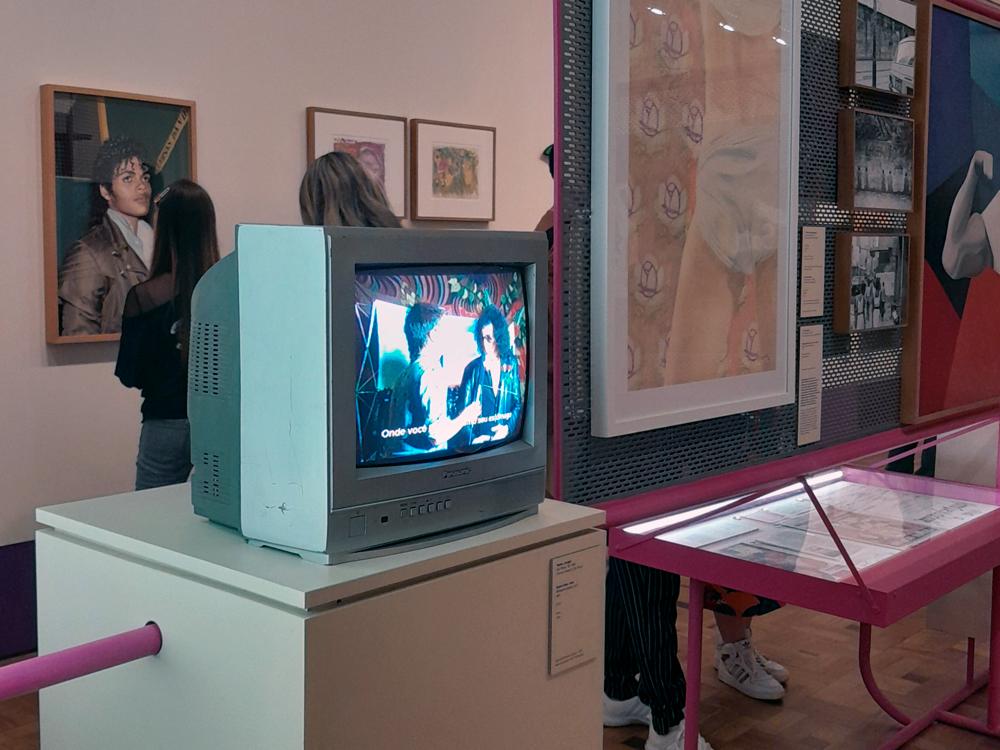 Fotografia de uma instalação da exposição Fullgás, exibindo um televisor de tubo Panasonic sobre pedestal branco com laterais pintadas de verde-claro. Na tela, uma cena de entrevista legendada. Ao fundo, obras penduradas na parede, incluindo um retrato de um cover de Michael Jackson e outras peças emolduradas. Há visitantes observando as obras.