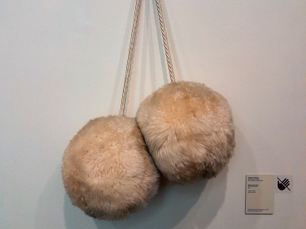 Fotografia de uma obra composta por duas esferas fofas revestidas de pele sintética bege, penduradas lado a lado por uma corda presa à parede branca. À direita, uma pequena placa com informações da peça.