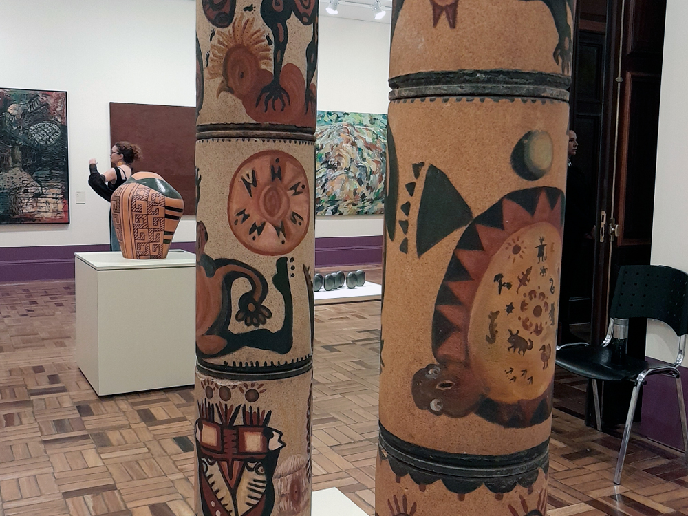 Detalhes de arte indígena em vasos de cerâmica expostos em Fullgás, com visitas observando as peças e obras no fundo.
