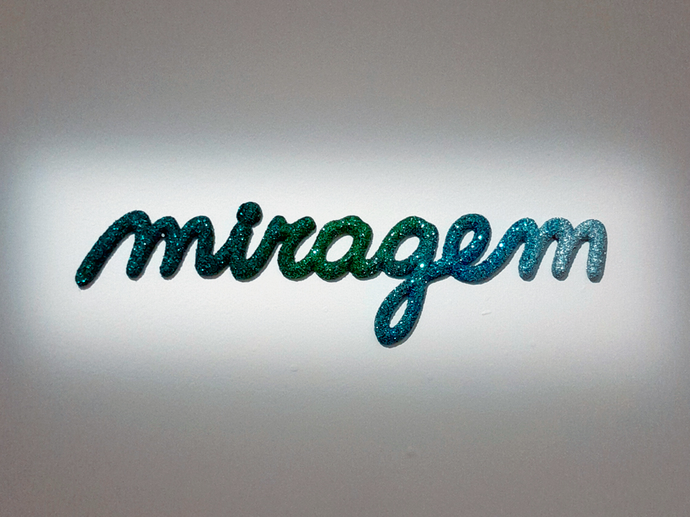 Escultura de parede com a palavra miragem escrita com letras brilhantes em um fundo branco, destacando-se pelo efeito de glitter colorido em tons de verde, turquesa e azul, formando degradê.