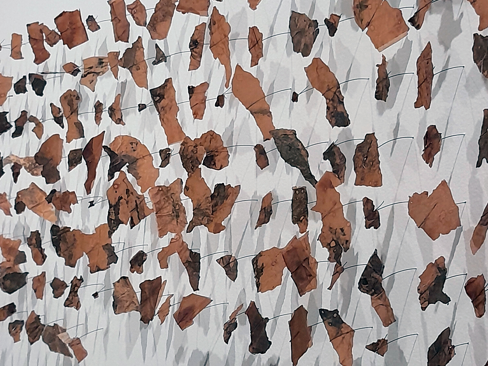 Imagem de arte contemporânea com folhas de papel em tons de marrom, preto e branco, criando uma textura abstrata e dinâmica na parede branca.