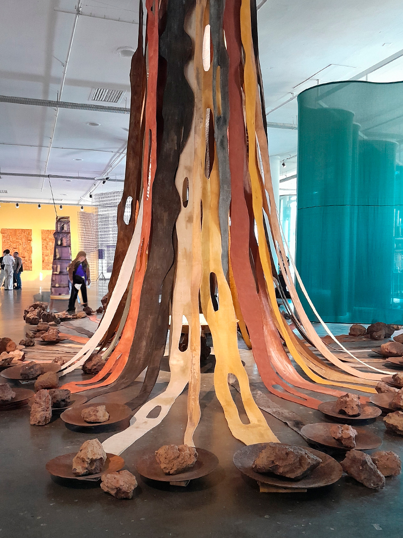 Instalação de grande escala composta por longas tiras verticais de tecido ou material maleável em tons terrosos — marrom, ocre, laranja, bege e vermelho — pendendo do teto e se espalhando pelo chão como raízes. Ao redor das tiras, há pedras dispostas sobre discos metálicos circulares, criando um conjunto que remete à força da natureza e à ancestralidade. O ambiente é amplo, com piso de cimento e divisórias translúcidas em verde-água ao fundo.