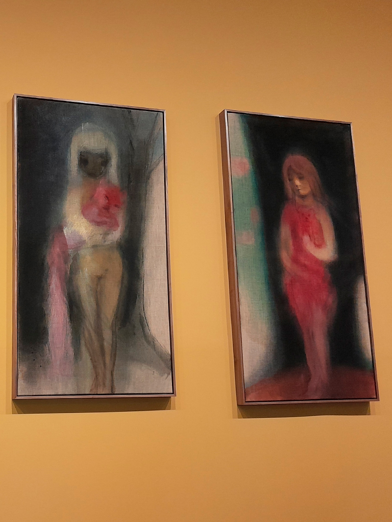 Duas pinturas verticais lado a lado, expostas em uma parede de tom ocre. Ambas retratam figuras femininas em traços difusos e cores suaves, com predominância de tons escuros e rosados. A figura da esquerda parece nua, com um pano rosa nas mãos; a da direita veste algo vermelho e encara o chão. As pinceladas e o efeito de desfoque criam uma atmosfera etérea e introspectiva.