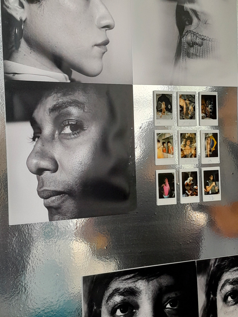 Composição fotográfica exibindo retratos em preto e branco de rostos humanos em close, intercalados com pequenas fotos coloridas no formato de Polaroid. Os rostos são mostrados de diferentes ângulos, destacando expressões e detalhes como olhos, pele e cabelos. As Polaroids menores mostram cenas casuais e pessoas em ambientes diversos. O fundo metálico reflete levemente a luz, adicionando textura à montagem.