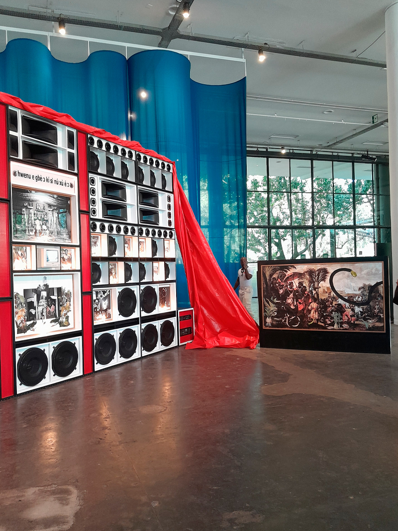 Instalação com uma grande estrutura retangular composta por caixas de som e painéis fotográficos, emoldurada por uma moldura vermelha e parcialmente coberta por um tecido de vinil vermelho. À direita, uma tela exibe uma colagem com figuras históricas e um enorme lagarto pintado sobre a cena. Ao fundo, cortinas de tecido azul translúcido descem do teto, contrastando com a luz natural que entra pelas janelas amplas do pavilhão.