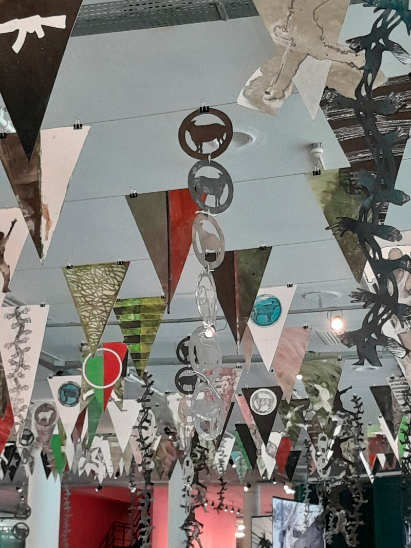 Conjunto de bandeirolas triangulares suspensas no teto, formando uma instalação aérea colorida. Cada bandeira apresenta desenhos e colagens diversas — armas, animais, formas abstratas e manchas de tinta — em tons de marrom, verde, preto, branco e vermelho. As bandeirolas estão presas por fios metálicos, criando um emaranhado visual que ocupa todo o espaço superior da sala.