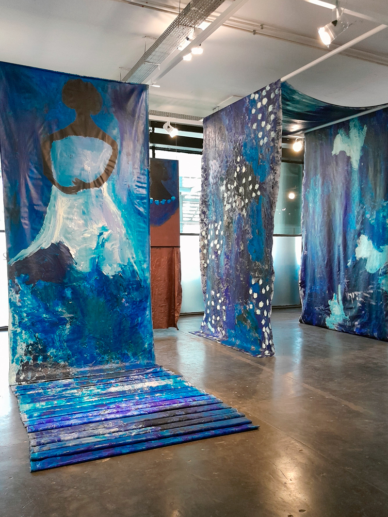 Grande instalação no pavilhão da Bienal, composta por tecidos pintados em tons de azul, pendurados verticalmente e estendidos pelo chão. As telas trazem figuras humanas estilizadas e formas orgânicas, com pinceladas expressivas e áreas pontilhadas de branco. A luz do teto reflete nos tecidos, criando brilhos e texturas que evocam movimento e profundidade.