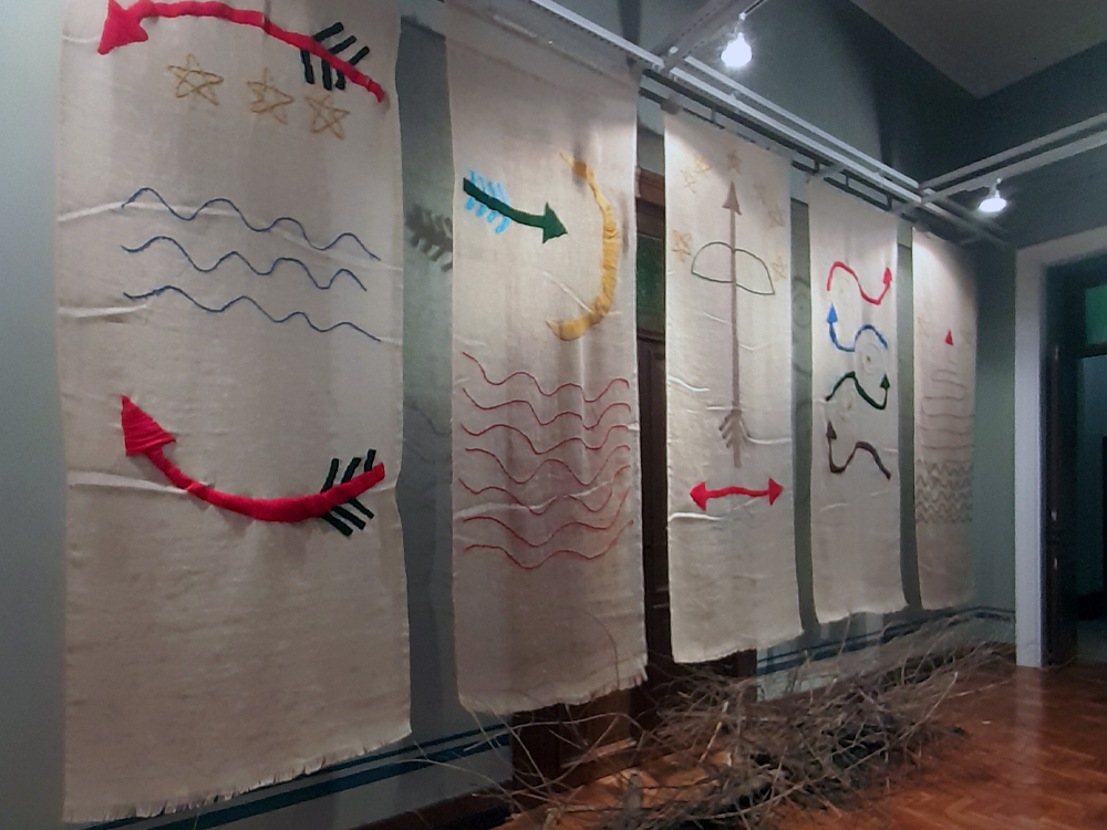 A imagem mostra uma instalação artística composta por cinco grandes painéis de tecido cru pendurados verticalmente, lado a lado, em uma parede cinza. Cada painel apresenta bordados coloridos com formas geométricas, setas e ondas, criando um visual simbólico e abstrato. No primeiro painel, à esquerda, há três estrelas bordadas em amarelo, linhas onduladas azuis e duas setas vermelhas com penas pretas nas extremidades. O segundo painel traz linhas onduladas vermelhas, uma seta verde apontando para a direita e uma meia-lua amarela. O terceiro painel exibe uma grande seta marrom vertical com um arco semicircular no topo e uma seta dupla horizontal vermelha na base. O quarto painel tem linhas onduladas coloridas (vermelho, azul, verde e marrom) e pequenas setas direcionais. O quinto e último painel mostra uma seta vermelha no topo e linhas onduladas cinza na parte inferior. No chão, na frente dos painéis, há um conjunto de galhos secos espalhados, adicionando textura à composição. A iluminação da sala é feita por spots de teto.