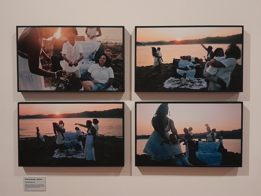 A imagem mostra um conjunto de quatro fotografias expostas lado a lado na parede, emolduradas em preto. As fotos retratam um grupo de pessoas negras, majoritariamente mulheres e crianças, reunidas à beira de um rio ou mar durante o pôr do sol. Todas usam roupas brancas, evocando referências a tradições afro-brasileiras e rituais de ancestralidade. Na fotografia superior esquerda, uma mulher segura um recipiente enquanto outra, sentada, sorri, e uma criança está entre elas. Na imagem superior direita, o grupo se posiciona diante da água, algumas pessoas seguram crianças e outras gesticulam, com o céu alaranjado ao fundo. A foto inferior esquerda mostra um enquadramento mais aberto, revelando a paisagem com o pôr do sol intenso, enquanto as pessoas interagem sobre uma toalha branca estendida no chão. Na fotografia inferior direita, o contra-luz acentua as silhuetas e as texturas das roupas, destacando a delicadeza do gesto de uma mulher que segura um bebê.
