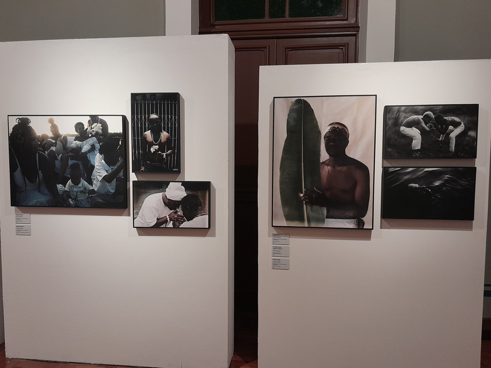A imagem mostra uma parede expositiva com duas seções de 3 fotografias cada. No conjunto da esquerda, a maior, na parte inferior, retrata várias pessoas negras, majoritariamente mulheres e crianças, reunidas, vestindo roupas brancas, evocando rituais de matriz africana. Acima, uma foto vertical mostra uma pessoa negra com colares e flores, posicionada diante de uma grade, olhando para baixo. Abaixo, outra imagem menor mostra duas pessoas, também de branco, tocando as testas uma na outra num gesto de afeto ou bênção. Já no conjunto da direita, a principal, maior e central, retrata uma pessoa negra de pele retinta, com o tronco nu e um pano branco na cabeça, segurando uma folha grande de bananeira, em um enquadramento delicado e contemplativo. Acima, uma fotografia em preto e branco mostra duas pessoas ajoelhadas frente a frente, em um gesto de conexão e devoção. Abaixo, outra imagem em preto e branco captura uma pessoa submersa na água, criando uma atmosfera silenciosa e introspectiva.