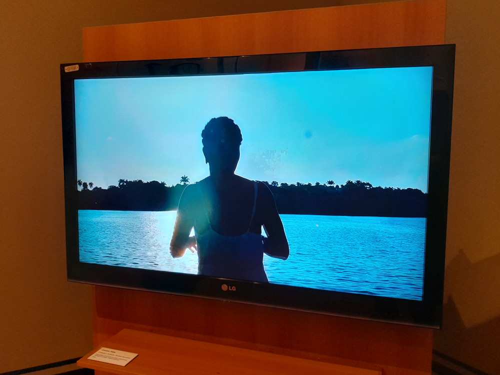 Tela de TV exibindo um vídeo da exposição O Som Que Ressoa em Mim Foi Meu Ancestral Que Tocou. A imagem apresenta a silhueta de uma pessoa de costas, em primeiro plano, usando uma blusa de alças brancas, diante de um rio iluminado pela luz do sol. A cena é capturada no momento em que o sol se põe ou nasce, criando um brilho azulado intenso que reflete na água e contrasta com a vegetação escura no horizonte.