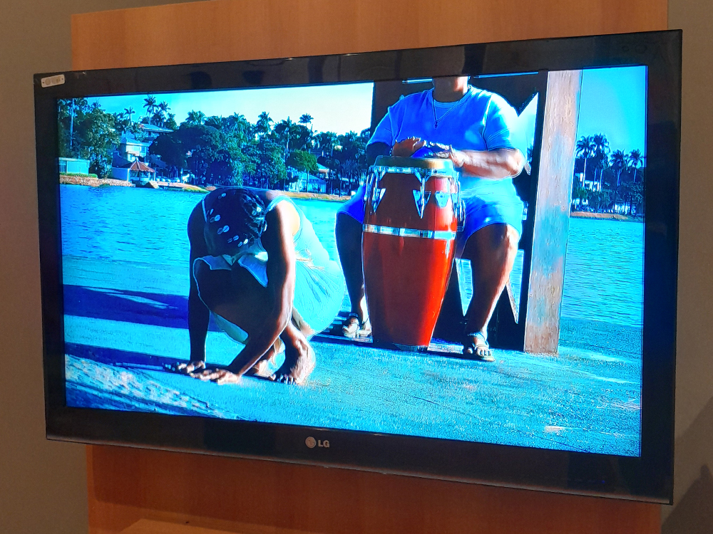 No centro de uma tela de TV, uma pessoa está ajoelhada e inclinada para frente, tocando o chão com as mãos, enquanto outra pessoa, parcialmente visível, está sentada atrás, tocando um tambor vermelho. A cena acontece em um píer de madeira sobre um rio, com casas, coqueiros e céu azul ao fundo. A iluminação é intensa e as cores são vibrantes.
