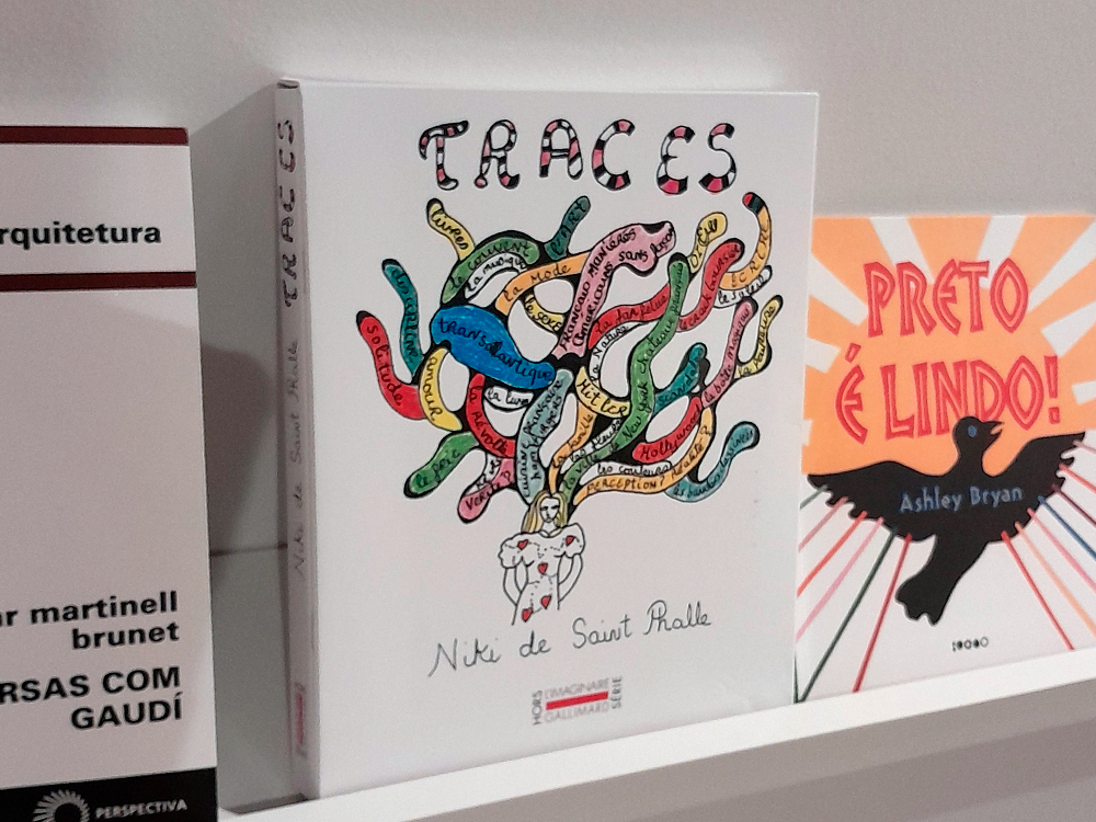 Fotografia de uma prateleira de livros exibindo o livro Traces, de Niki de Saint Phalle, entre outras obras. A capa branca mostra uma ilustração colorida de formas orgânicas entrelaçadas, com palavras escritas em francês dentro delas, e o título TRACES no topo em letras decoradas. À direita está o livro Preto é lindo! de Ashley Bryan, com fundo laranja e um pássaro preto; à esquerda, parte da lombada de outro livro sobre arquitetura.