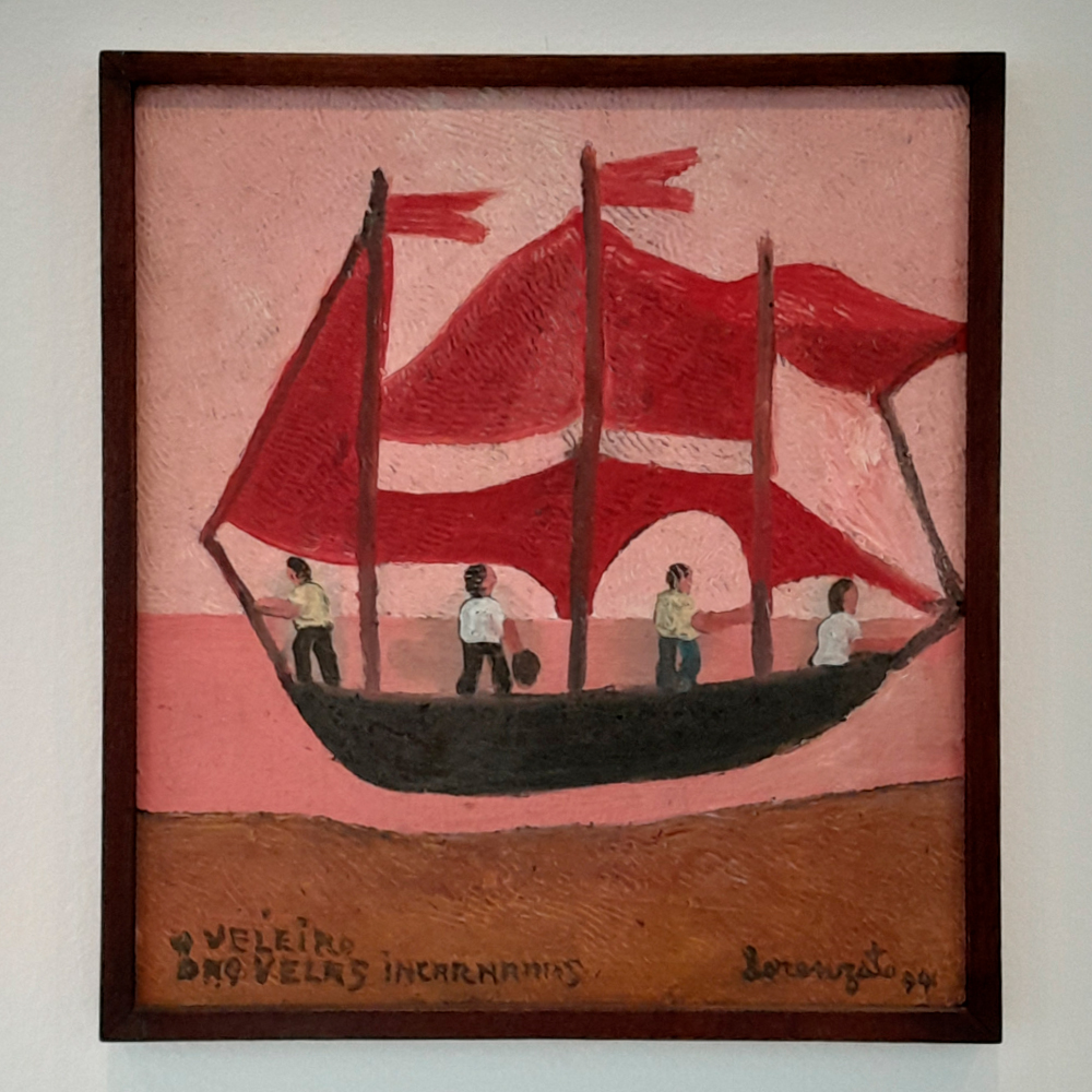 Pintura de Amadeo Luciano Lorenzato intitulada Veleiro das velas encarnadas, de 1994. A obra mostra um barco escuro navegando sobre um mar rosado, com quatro figuras humanas a bordo. As velas grandes e onduladas são vermelhas, com faixas claras, contrastando com o fundo rosa. A pintura, de textura espessa e traços simples, está emoldurada em madeira.