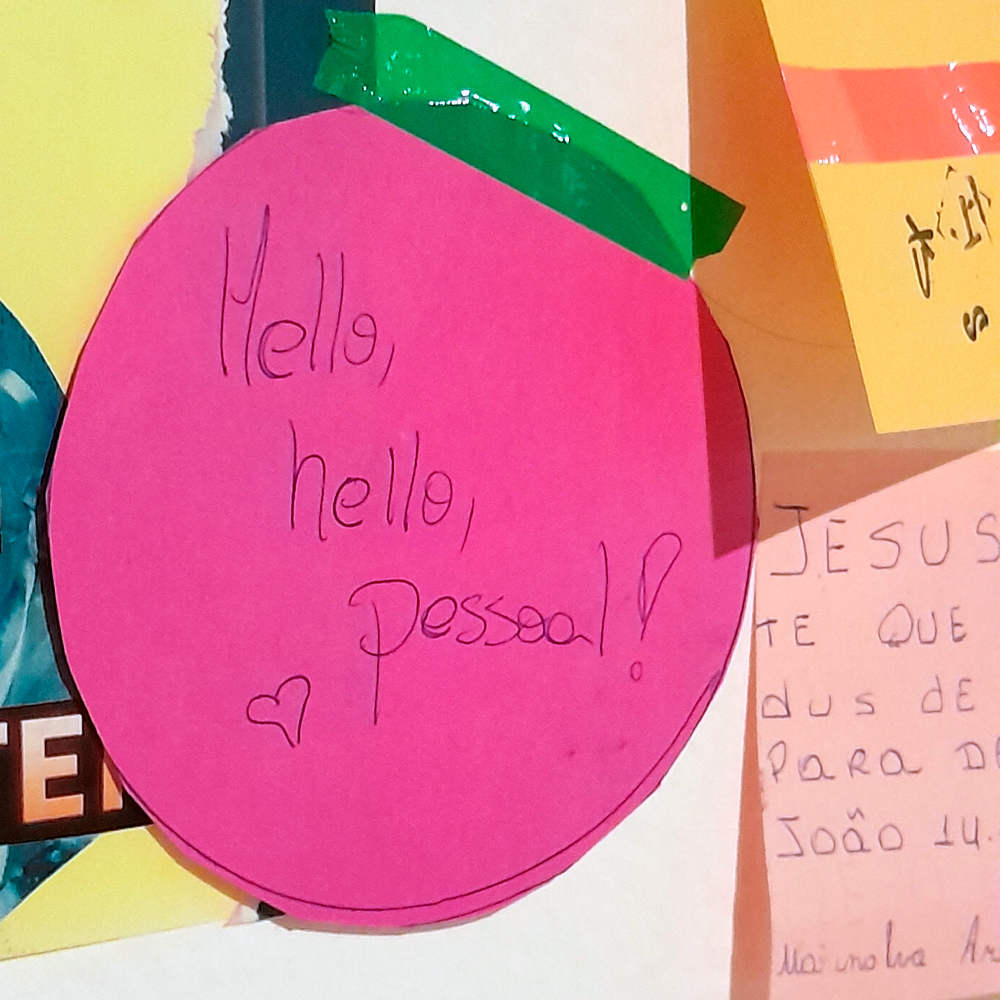 Recorte circular de papel rosa preso na parede com fita adesiva verde. Nele está escrito à mão, em caneta preta: Hello, hello, pessoal! seguido de um pequeno coração desenhado. Ao redor, aparecem outros papéis coloridos com escritos diversos parcialmente visíveis.