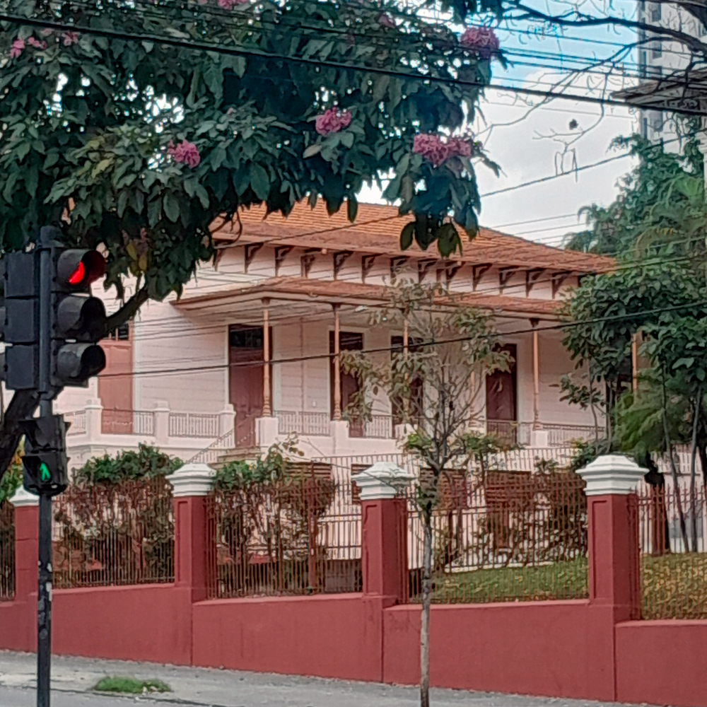 Fachada de um casarão antigo pintado em tons claros. O imóvel é cercado por um muro vermelho com grades metálicas e vegetação ao redor. Em primeiro plano, há um semáforo com luz vermelha acesa e uma árvore florida pendendo sobre a cena.