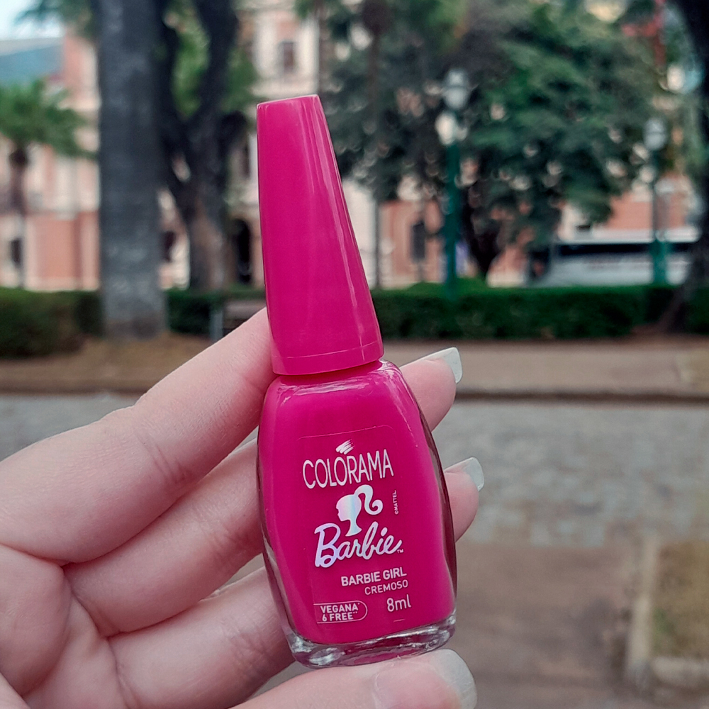 Mão segurando um frasco de esmalte rosa-choque da linha Colorama Barbie, na cor “ Barbie Girl, com o rótulo escrito vegana & free. Ao fundo, o cenário urbano mostra uma praça arborizada com árvores e construções históricas desfocadas.