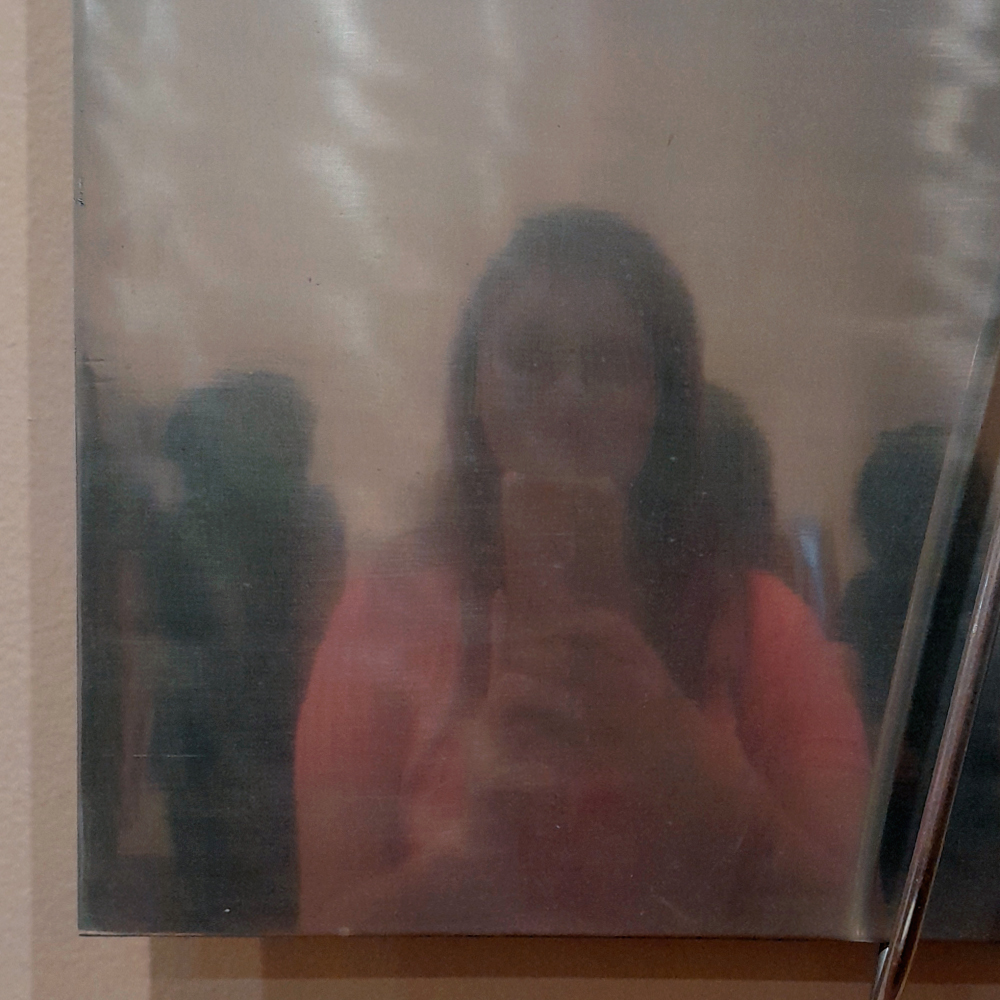 Reflexo de uma pessoa fotografando em uma superfície metálica polida e ligeiramente fosca, com aparência espelhada. A figura aparece desfocada, segurando o celular diante do rosto. Ao fundo, é possível ver outras silhuetas refletidas de pessoas no ambiente expositivo.