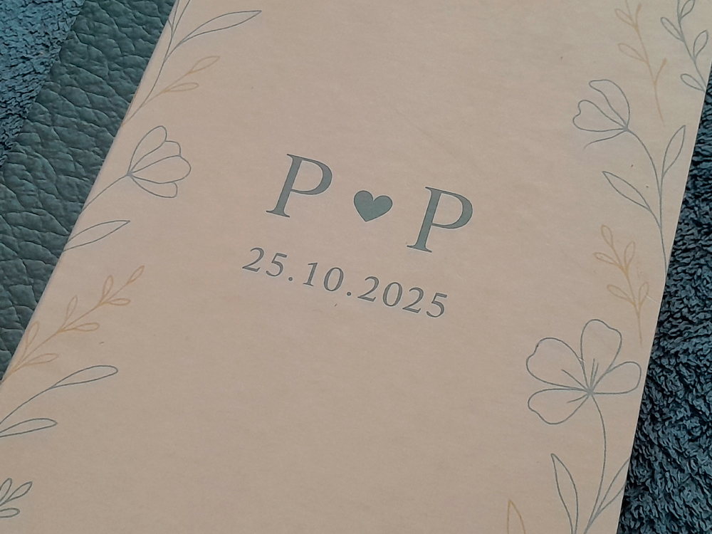 Foto de um caderno para o casamento com capa bege decorada por ilustrações delicadas de flores. No centro, as iniciais P ? P e a data 25.10.2025 impressas em azul acinzentado.