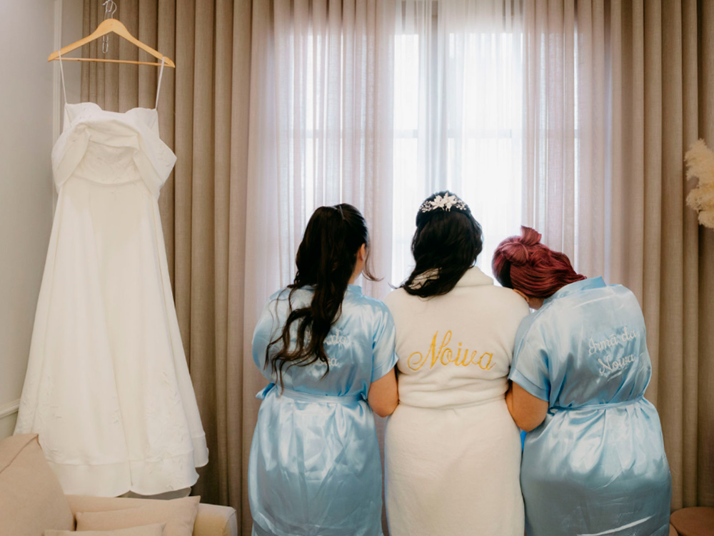Foto do making of do casamento. A noiva, de roupão branco bordado com a palavra Noiva, está de costas entre duas madrinhas. À esquerda, a madrinha Dani de cabelo preto comprido; à direita, a madrinha Luly, com cabelo rosa, ambas usando robes azul-claro bordados escritos Irmã da Noiva. À esquerda da cena, o vestido de noiva pendurado.