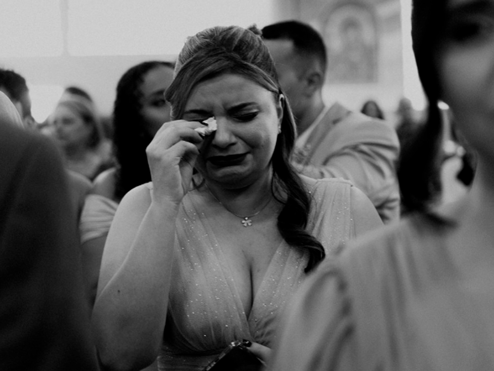 Foto em preto e branco da madrinha Luly emocionada durante a cerimônia de casamento. Ela enxuga as lágrimas com um lenço enquanto usa um vestido longo claro com brilho. Pessoas desfocadas ao fundo completam a cena.