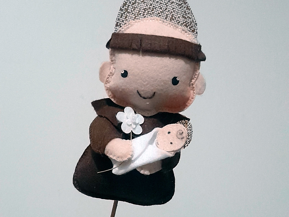 Foto de um topper artesanal de feltro representando Santo Antônio segurando o Menino Jesus. A figura é feita em tons de marrom e bege, com bochechas rosadas e expressão sorridente.