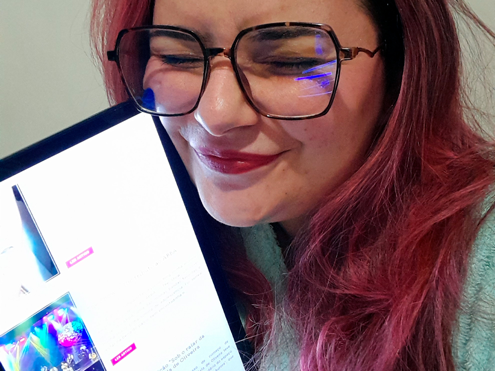 Fotografia da mesma mulher de cabelos rosa-escuros, agora sorrindo com os olhos fechados e o rosto encostado carinhosamente no tablet que segura, onde o blog está aberto na mesma página.