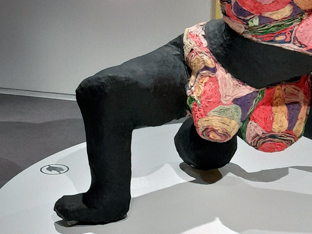 Detalhe de uma escultura de Niki de Saint Phalle mostrando parte das pernas e quadris de uma figura feminina robusta. A pele é preta e a roupa, feita com texturas e cores vibrantes, lembra tecidos enrolados em tons de rosa, roxo, verde e amarelo. A escultura está sobre uma base branca de museu, com o símbolo de “não tocar” visível no chão.