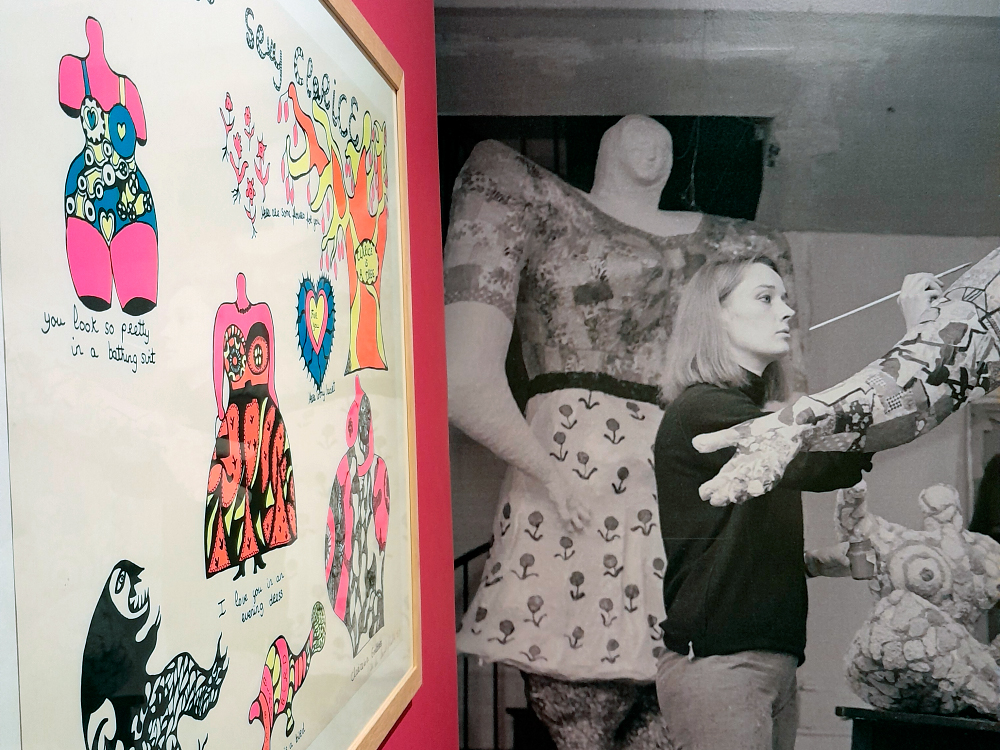 Fotografia mostrando uma parede expositiva com dois painéis lado a lado. À esquerda, ilustrações coloridas de figuras femininas estilizadas, em traços característicos de Niki de Saint-Phalle, com cores intensas como rosa, azul e laranja. À direita, uma fotografia em preto e branco da artista pintando uma de suas grandes esculturas “Nana”. Ela segura um pincel e trabalha concentrada diante de uma figura feminina monumental.