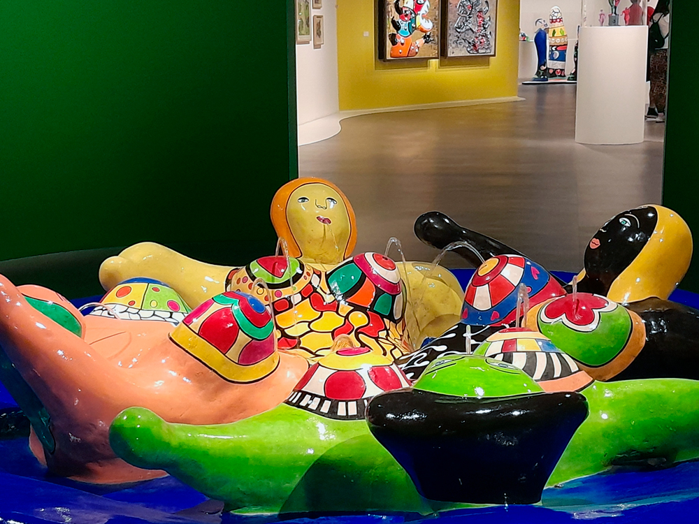 Escultura de grandes proporções composta por figuras femininas coloridas, de formas arredondadas e alegres, dispostas em círculo sobre uma base azul que funciona como fonte, com jatos de água saindo das esculturas. As figuras são pintadas com padrões vibrantes em vermelho, verde, amarelo e preto. Ao fundo, é possível ver parte da exposição com paredes verde e amarela e outras obras de Niki de Saint Phalle expostas.