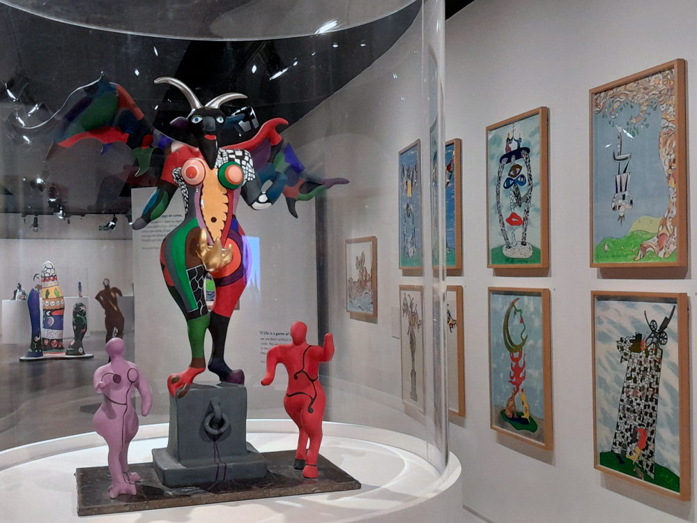 No centro da imagem, protegida por uma vitrine, encontramos uma escultura representando o arcano do Diabo no tarot com cores vibrantes e formas arredondadas. Ao fundo, dispostos na parede, vemos uma série de pinturas emolduradas que parecem explorar temas semelhantes: figuras estilizadas, cores vivas e uma estética que mistura o lúdico com o simbólico.