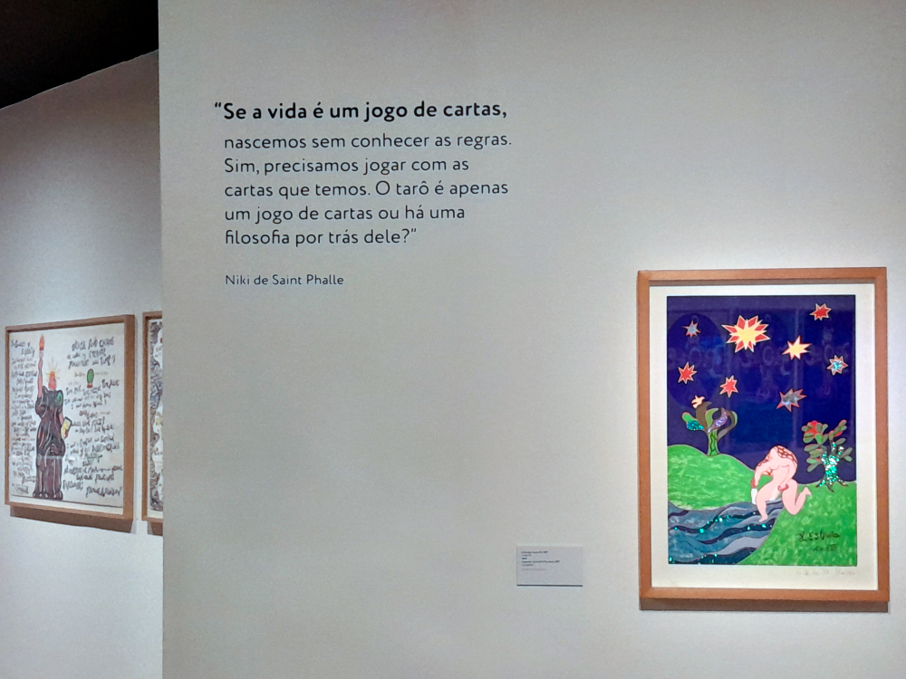 Imagem de uma parede de galeria de arte com quadro colorido do arcano da estrela, representado por uma mulher nua com 7 estrelas no céu escuro. Ao seu lado há a citação SE A VIDA É UM JOGO DE CARTAS, NASCEMOS SEM CONHECER AS REGRAS. SIM, PRECISAMOS JOGAR COM AS CARTAS QUE TEMOS, O TAROT É APENAS UM JOGO DE CARTAS OU HÁ UMA FILOSOFIA POR TRÁS DELE?, de Niki de Saint Phalle