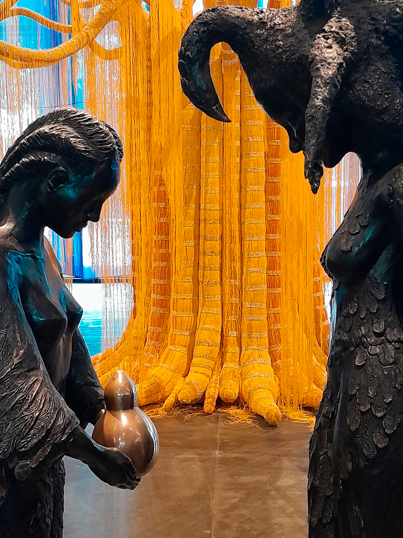 Duas esculturas em bronze dialogam frente a frente — à esquerda, uma figura feminina com traços humanos segura uma cabaça metálica; à direita, uma figura com corpo emplumado e cabeça de carneiro. Atrás delas, uma grande instalação feita de fios grossos amarelos desce do teto como raízes luminosas, preenchendo o espaço e trazendo textura vibrante à cena.
