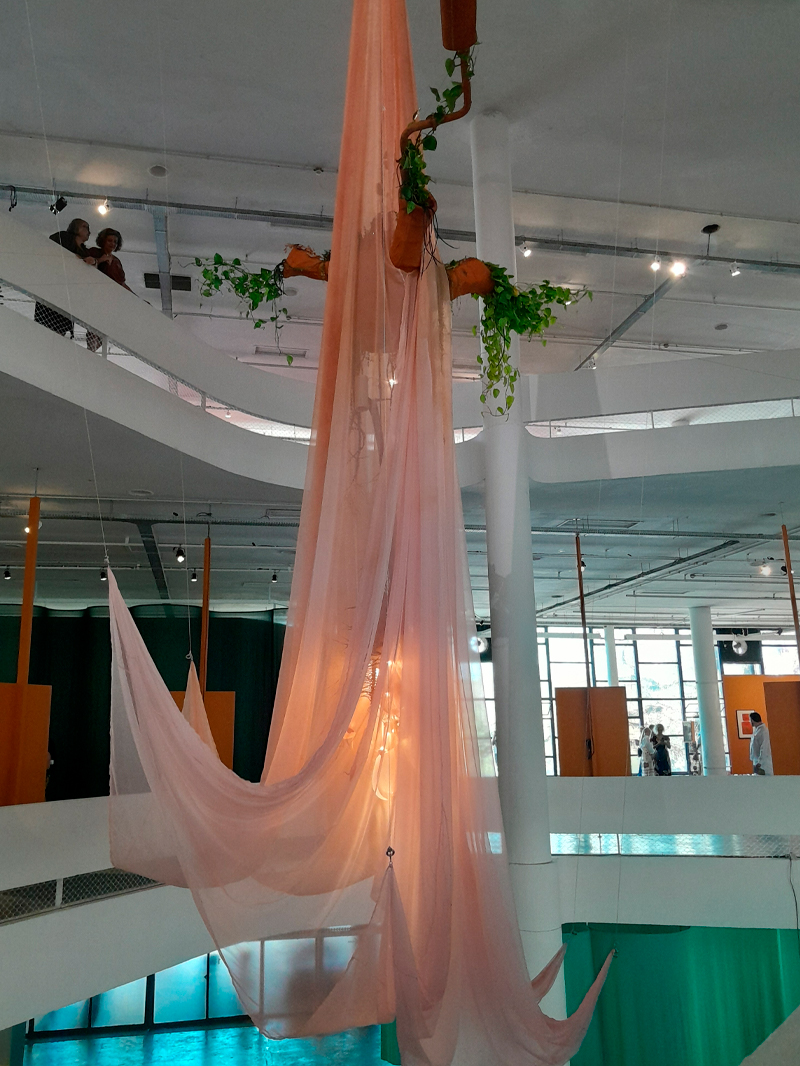 Instalação suspensa no vão livre do Pavilhão da Bienal, composta por tecidos translúcidos cor-de-rosa pendendo do teto em ondas, entrelaçados a estruturas de cobre e plantas vivas. A luz natural atravessa as camadas de tule e cria um efeito etéreo. No fundo, o espaço modernista do edifício com mezaninos brancos e visitantes observando as obras.