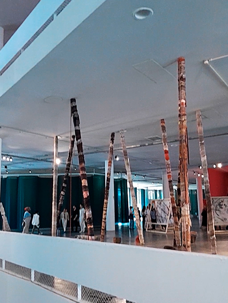 Vista de uma instalação composta por longos troncos verticais apoiados no chão e no teto do prédio da Bienal de São Paulo. As peças, cobertas por tecidos ou fibras em tons terrosos e esbranquiçados, formam uma espécie de floresta fragmentada dentro do espaço expositivo. Ao fundo, pessoas caminham entre as obras, cercadas por paredes verdes e luz suave.