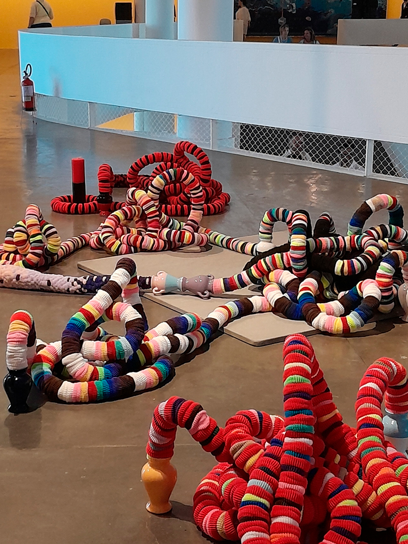 Instalação formada por grandes estruturas sinuosas, cobertas por crochês coloridos em listras vibrantes de vermelho, azul, verde, amarelo e branco. As formas lembram serpentes ou tentáculos espalhados pelo chão do pavilhão, criando um ambiente lúdico e têxtil. Ao fundo, o corrimão branco e as colunas do edifício complementam a cena.