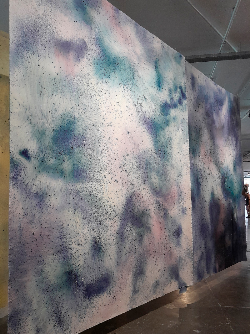 Painéis verticais de grandes dimensões exibem superfícies pintadas com manchas e respingos em tons de azul, lilás e rosa claro, formando uma textura etérea e difusa. A obra ocupa o espaço central da galeria da Bienal.