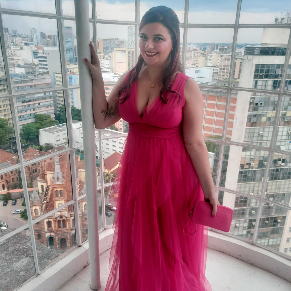 Luly posando em pé diante de uma grande janela envidraçada que mostra prédios altos e uma igreja antiga vista de cima. Ela usa um vestido longo rosa-pink de tule, com decote em V e saia esvoaçante em camadas. O cabelo está meio-preso, com ondas soltas e mechas rosadas. Na mão direita, ela segura uma clutch rosa que combina com o vestido. A luz natural realça a textura do tecido e o fundo urbano.