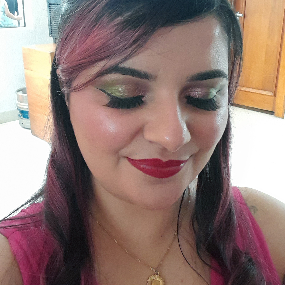 Close do rosto de Luly com os olhos fechados, mostrando uma maquiagem colorida e brilhante: sombra verde cintilante no canto externo e rosado canto interno, com essas cores se encontrando ao centro, com delineado preto bem marcado e cílios volumosos. Ela usa batom vermelho intenso e blush iluminado. O cabelo aparece com mechas rosadas e onduladas, e ela veste o mesmo vestido rosa.
