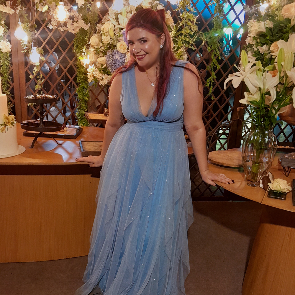 Luly posa sorrindo diante de uma mesa de madeira decorada para festa, cercada por arranjos de flores brancas, verdes e azuis, além de luzes amareladas penduradas que criam um clima aconchegante. Ela usa um vestido longo azul-claro de tule com brilho discreto, decote em V profundo e saia esvoaçante em camadas. O cabelo está meio-preso, com ondas e mechas rosadas contrastando com o azul do vestido. Ela apoia as mãos na mesa, inclinando levemente o corpo para a frente, com expressão alegre e confiante.