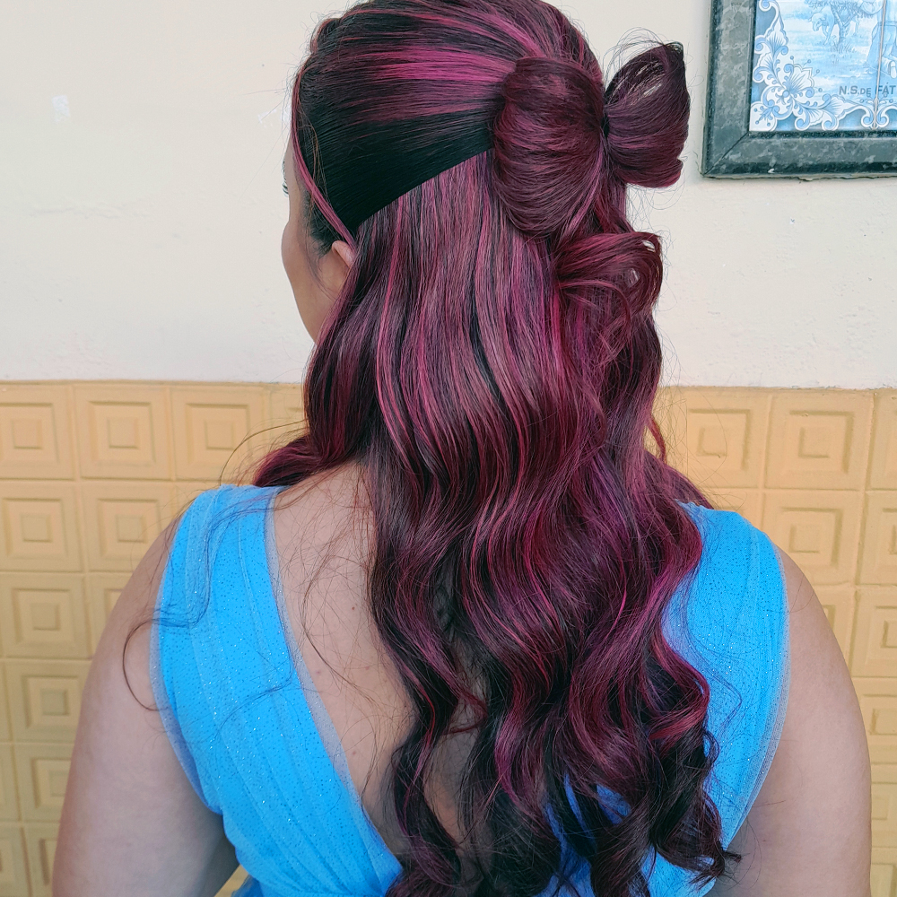 Mulher vista de costas, com os cabelos longos e ondulados em tons de rosa magenta e preto. O penteado é meio preso, com a parte superior formando um laço de cabelo estruturado, deixando as ondas soltas caindo pelas costas. A cor fantasia destaca mechas vibrantes e brilhosas. Ela veste um vestido azul claro com brilho discreto. Ao fundo há uma parede com revestimento amarelo geométrico e parte de um quadro pendurado.