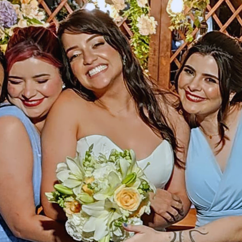Três mulheres abraçadas e sorrindo em um ambiente de festa decorado com flores claras e luzes quentes. Ao centro está a noiva, usando um vestido branco tomara que caia e segurando um buquê com flores brancas e amarelas. À esquerda e à direita, duas madrinhas vestindo vestidos azul-bebê; ambas têm maquiagem marcada com batom vermelho. A madrinha da esquerda tem cabelo preso com mechas rosadas, e a da direita usa um penteado baixo. Todas estão sorrindo com muita alegria, transmitindo clima de celebração.